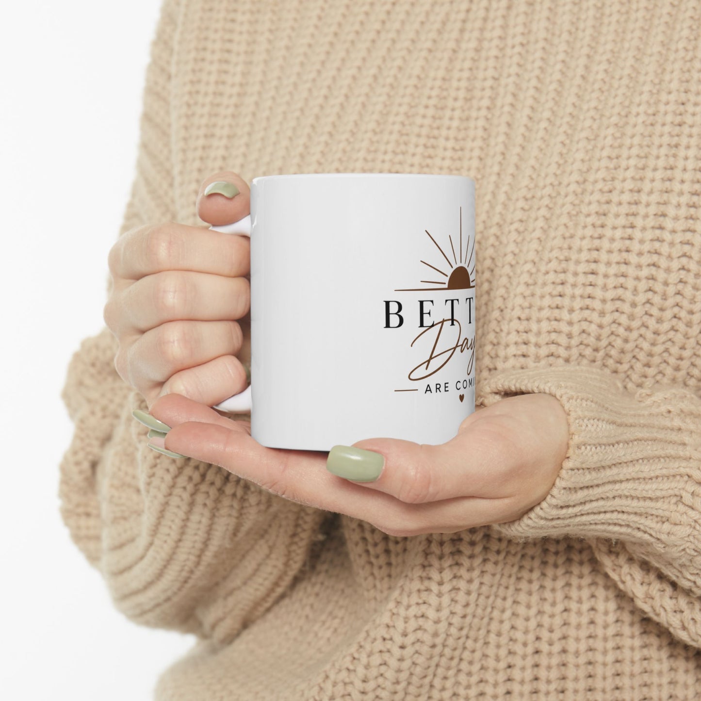 Person holding white coffee mug with Better Days Are Coming sunrise and heart inspirational design in natural light