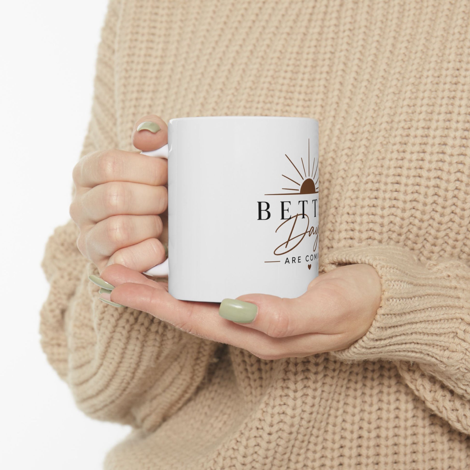 Person holding white coffee mug with Better Days Are Coming sunrise and heart inspirational design in natural light
