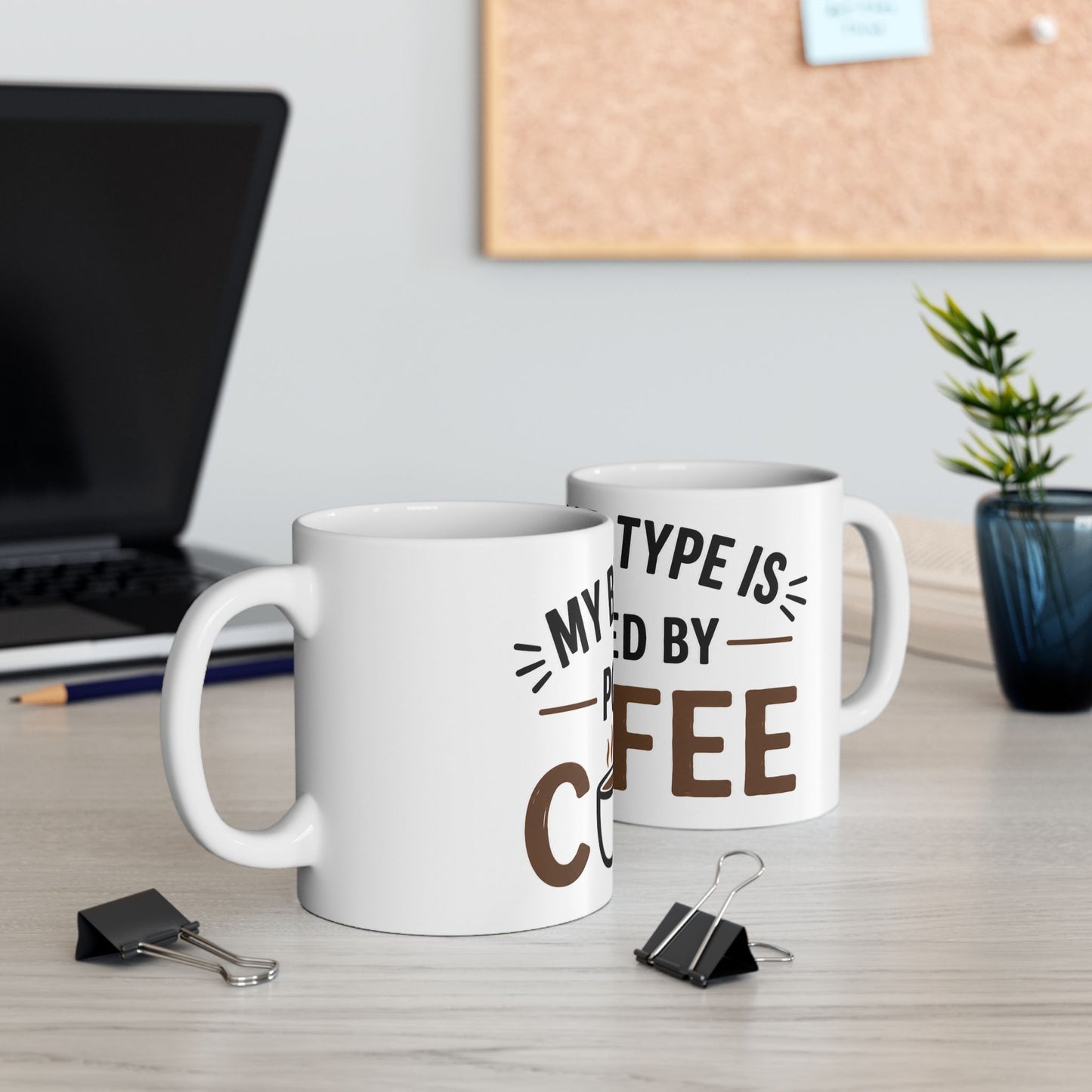 White coffee mug on desk next to computer showing My Blood Type Is Powered By Coffee funny coffee design in workspace setting